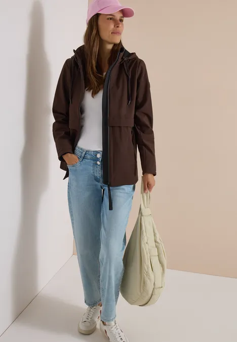 Jeune femme en veste marron, t-shirt blanc, jean bleu clair, casquette rose et grand sac en toile.