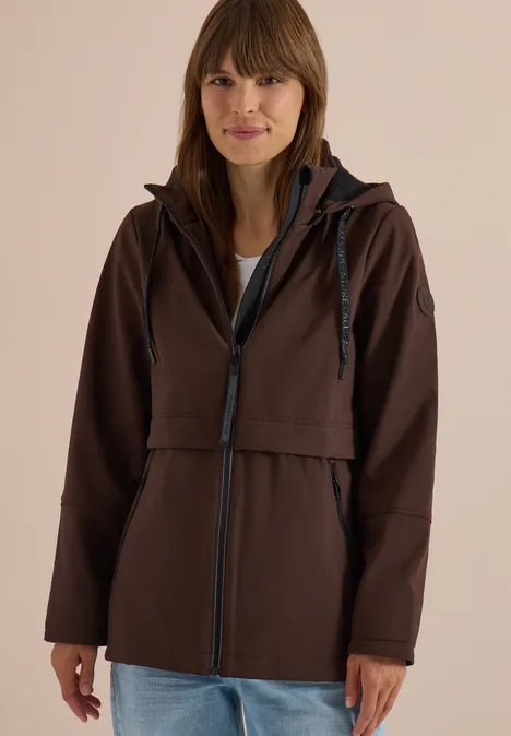 Femme portant une veste à capuche marron, posant souriante devant un fond neutre.