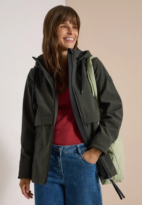 Softshell Jacke mit abnehmbarer Kapuze olive night Frau in dunkelgrünem Outdoor-Jacke, rotem Pullover und Jeans, lächelnd mit Rucksack auf der Schulter.