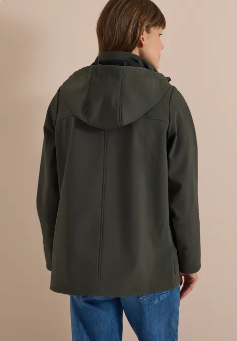Softshell Jacke mit abnehmbarer Kapuze olive night Eine Person von hinten in dunkelgrüner Kapuzenjacke und Jeans vor beiger Wand.