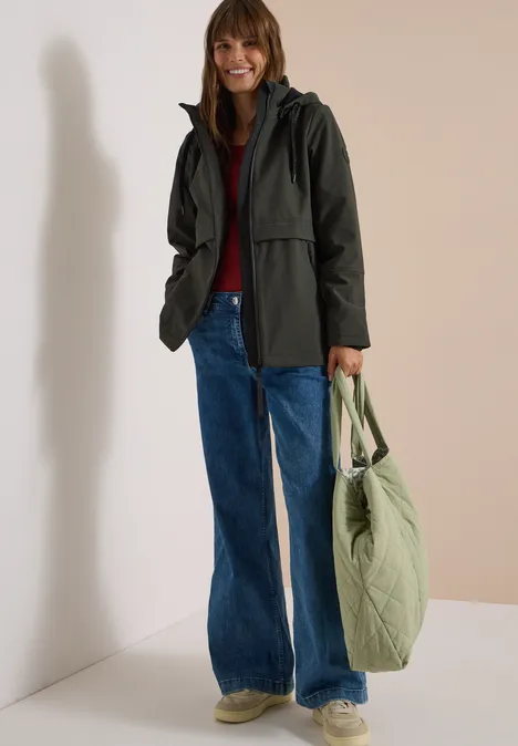 Softshell Jacke mit abnehmbarer Kapuze olive night Frau in grüner Jacke, rotem Pullover, Jeans und Sneakern, hält grüne Stepp-Tasche vor neutralem Hintergrund.