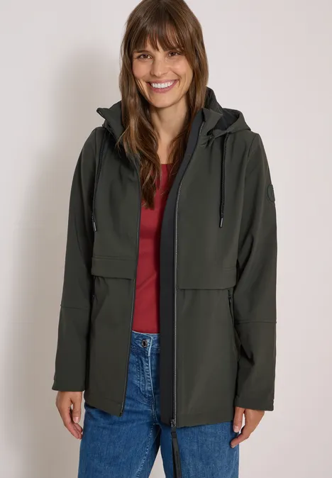 Softshell Jacke mit abnehmbarer Kapuze olive night Eine Frau lächelt und trägt eine grüne Jacke über einem roten Shirt und Jeans vor neutralem Hintergrund
