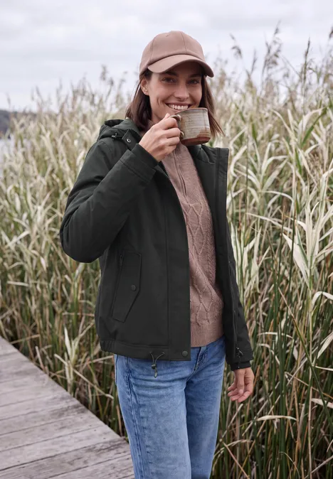 Een vrouw met jas, spijkerbroek en pet drinkt koffie uit een kopje op een steiger voor hoog riet.