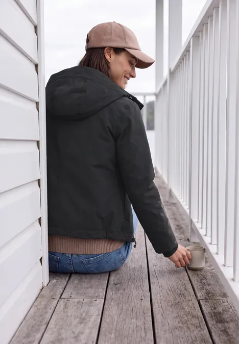 Vrouw met zwarte jas, blauwe jeans en pet zit op een houten terras, leunt op het balkon en glimlacht.