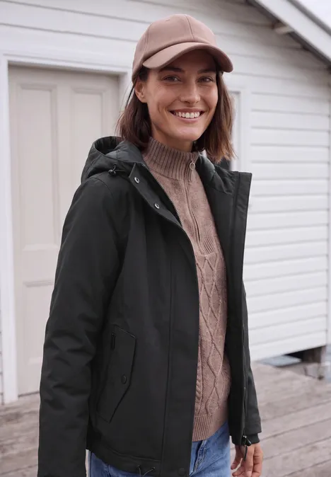 Vrouw met pet, gebreide trui en zwarte jas glimlacht voor een licht huis.