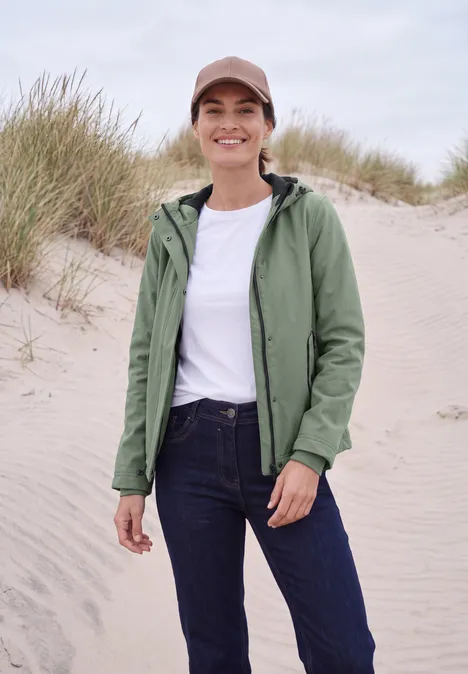 Een lachende vrouw in wit shirt, groene jas, spijkerbroek en pet op een zandstrand met duinen.