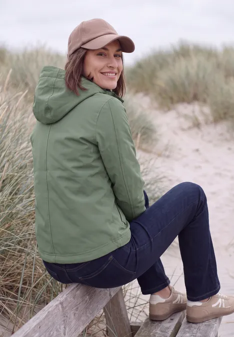 Vrouw in groen jack en spijkerbroek zit op een houten balk bij de duinen en kijkt glimlachend om zich heen.