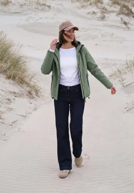Een vrouw in groene jas, spijkerbroek en pet loopt over zandduinen.
