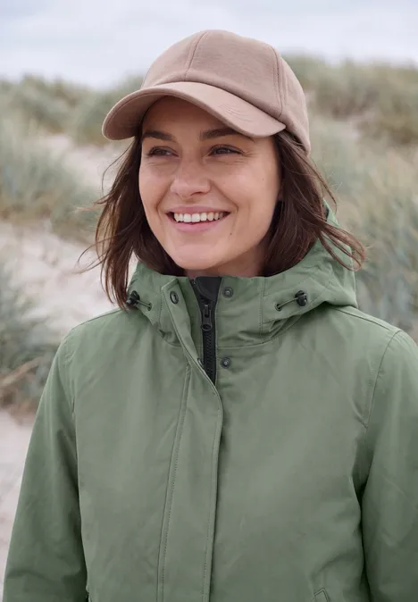 Glimlachende vrouw met groene jas en beige pet voor duinen.