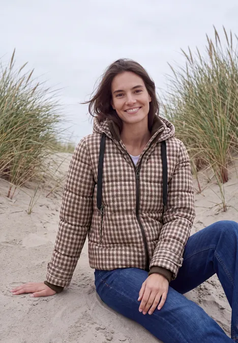 Jonge vrouw lacht in een geruite jas en spijkerbroek, zittend op duinen met helmgras.