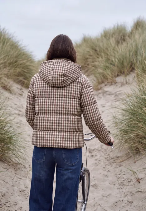 Een persoon in een geruite donsjas duwt een fiets tussen grasrijke duinen op het strand.