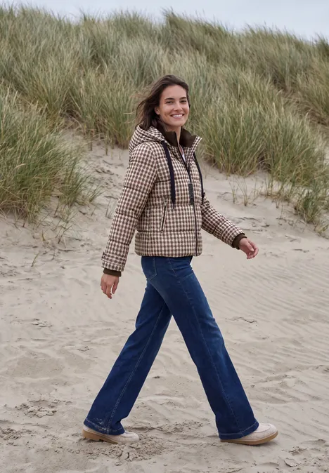 Een vrouw in geruite jas en flared jeans wandelt ontspannen over grasrijke duinen.