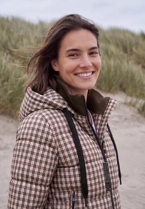 Een glimlachende jonge vrouw in een geruit jack staat voor duinen op het strand.