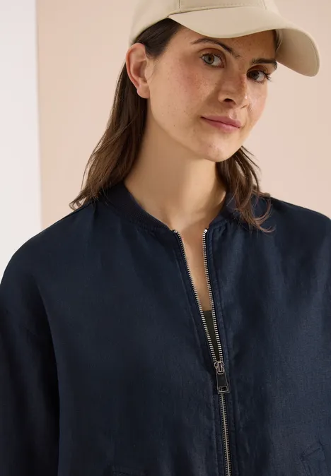 Eine Person in blauer Bomberjacke, hellem Top und beiger Baseballcap vor neutralem Hintergrund