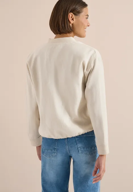 Persoon van achteren met een lichte trui en blauwe jeans tegen een neutrale achtergrond.