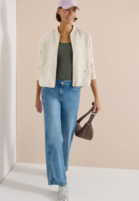 Vrouw in witte bomberjas, groen topje, blauwe wijde jeans, sneakers en een kleine bruine tas.