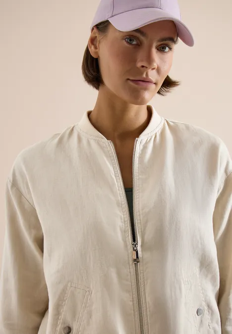 Vrouw in lichtbeige jas met ritssluiting en roze pet, voor een neutrale achtergrond.