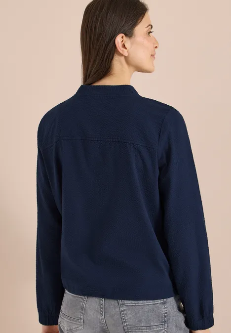 Rückansicht einer Frau in dunkelblauem Pullover und grauer Jeans vor neutralem Hintergrund.