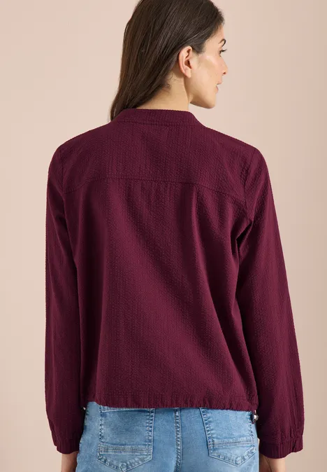 Blouson in Seersuckerqualität mit Zipper sweet grape red Frau von hinten in bordeauxroter Bluse und blauer Jeans vor beigem Hintergrund.