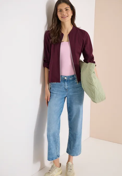 Blouson in Seersuckerqualität mit Zipper sweet grape red Eine Person lehnt an der Wand, trägt eine lila Jacke, ein rosa Top, blaue Jeans, weiße Sneaker und hält eine grüne Tasche.