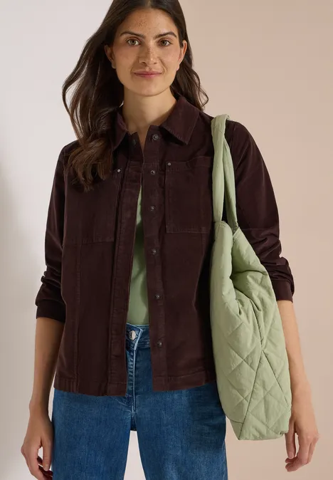 Eine Frau trägt eine braune Cordjacke, grüne Bluse, blaue Jeans und eine große, gepolsterte, grüne Tasche.