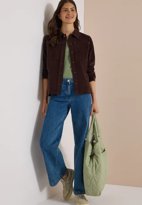 Frau trägt lockere Jeans, grünes T-Shirt, braune Cordjacke und hält eine große gesteppte grüne Tasche.