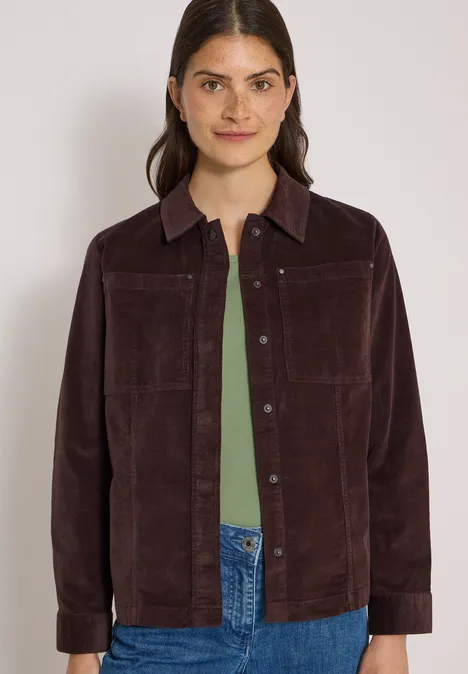 Junge Frau in brauner Cordjacke, grünem Top und Jeans, lächelt leicht vor neutralem Hintergrund.