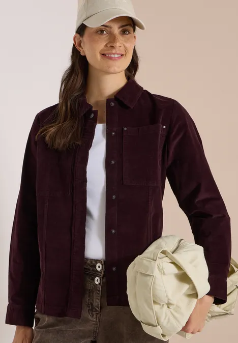 Frau in bordeauxfarbener Cordjacke, weißem T-Shirt und Cargohose hält lässig eine cremefarbene Tasche.