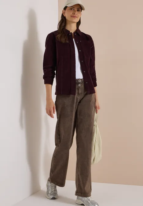 Frau im lässigen Look: weißes Top, bordeauxfarbene Jacke, braune Cordhose, silberne Sneaker, Cap.