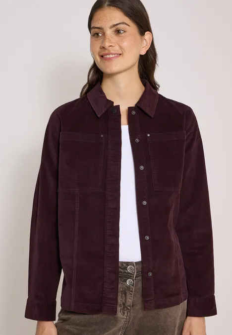 Eine junge Frau trägt eine dunkelviolette Kordjacke über weißem Shirt und gemusterten Hosen.