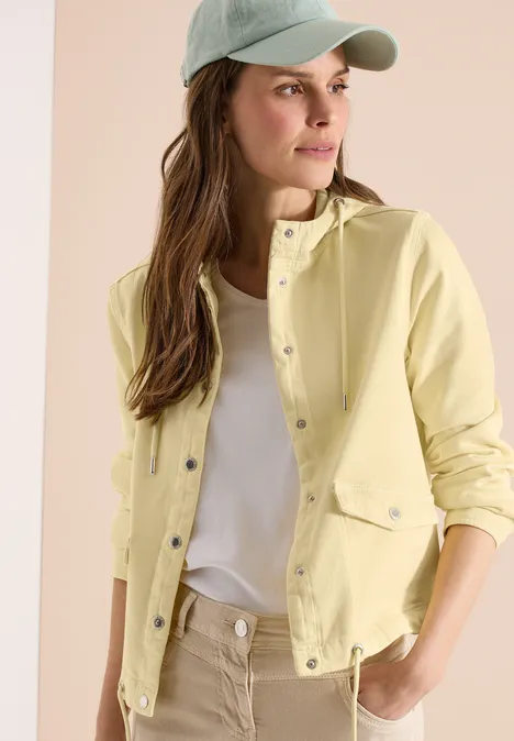 Jonge vrouw met gele jas, wit shirt en pet tegen een neutrale achtergrond.