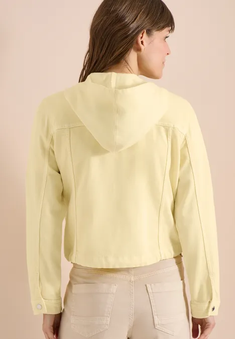 Vrouw van achteren in lichtgele capuchonblouse en beige broek.