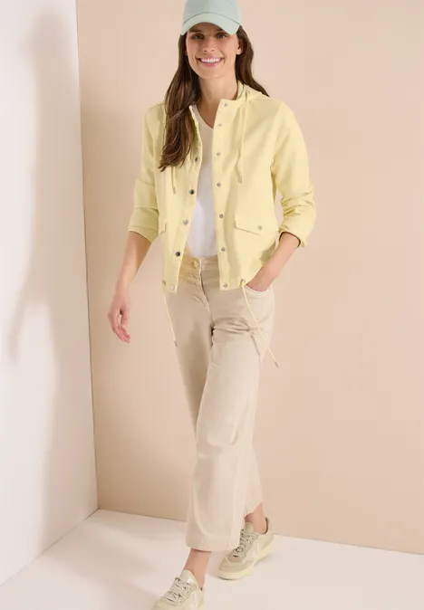 Vrouw staat in lichte ruimte, draagt gele jas, wit topje, beige broek en sneakers.