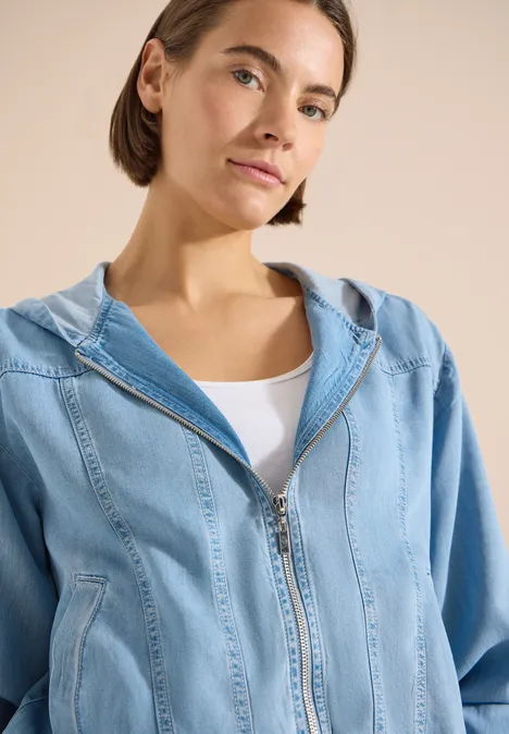Vrouw draagt lichtblauwe spijkerbroek-hoodie met rits en wit topje, gezicht onscherp gemaakt.