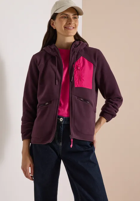 Frau trägt lila Kapuzenjacke mit pinker Brusttasche, Jeans und weiße Cap, lächelnd in Studio.