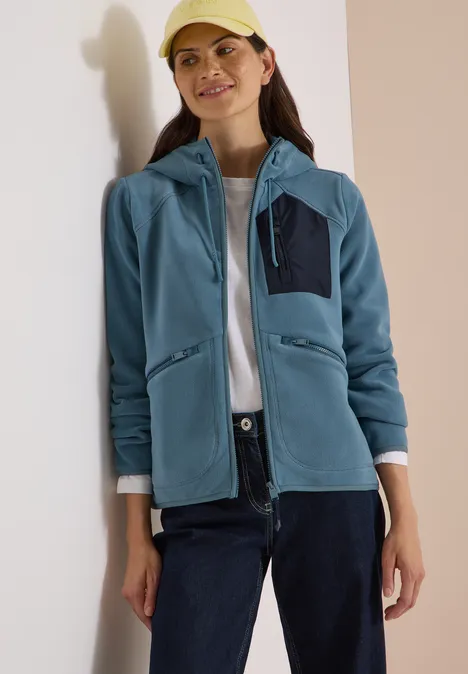 Frau in blauem Fleece-Hoodie, weißem Shirt, dunkler Hose und Cap vor neutralem Hintergrund.