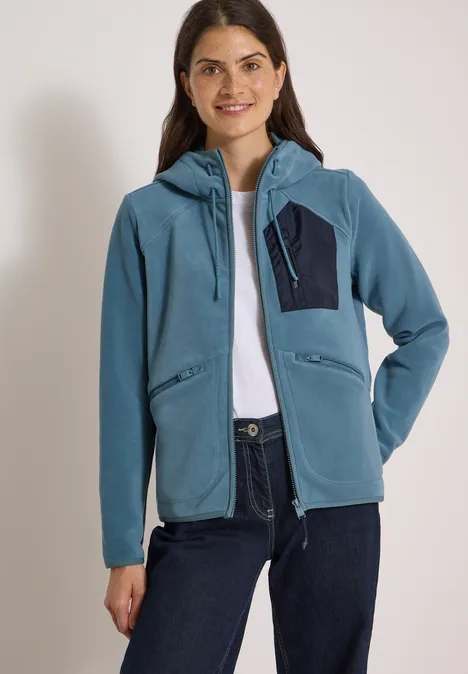 Eine lächelnde Frau trägt eine blaue Kapuzenjacke mit Reißverschluss und Brusttasche sowie eine Jeans.