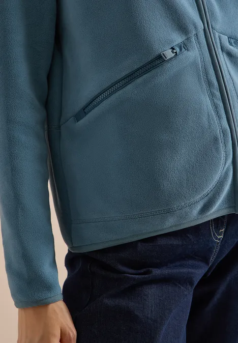 Person in türkisfarbener Fleecejacke mit Reißverschlusstasche und blauen Jeans.