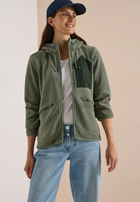 Frau trägt grüne Fleecejacke mit Kapuze, weiße Bluse, Jeans und Baseballcap.