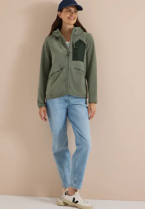 Frau in grüner Jacke, Jeans, Sneakern und Cap vor beigem Hintergrund.