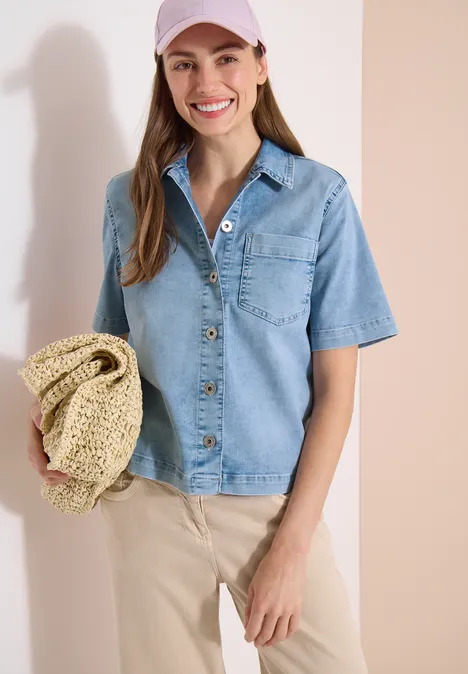 Frau in kurzärmeliger Jeansbluse, heller Hose hält Strohhut vor neutralem Hintergrund.