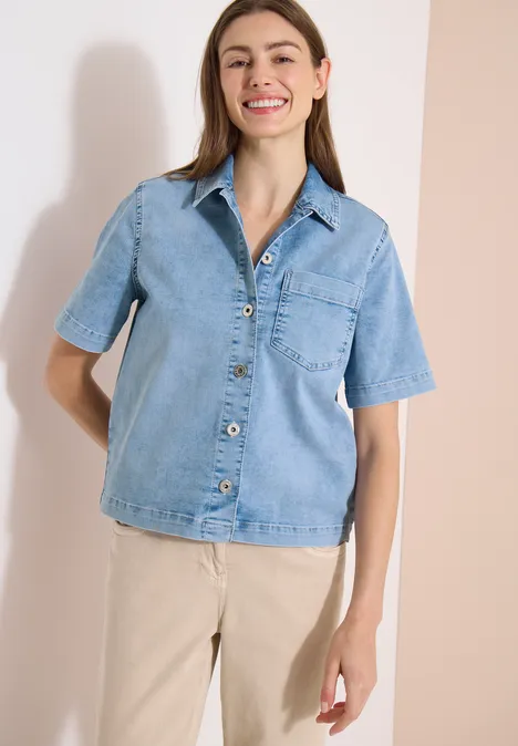 Frau in hellblauer Jeansbluse mit Knöpfen trägt beige Hose vor neutralem Hintergrund.