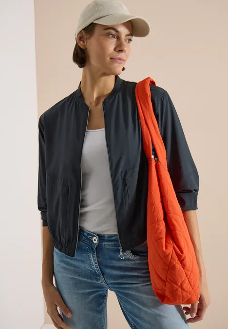 Blouson in Unifarbe mit Zipper palm breeze green Frau in Jeans, weißem Tanktop, schwarzer Jacke, beige Cap und oranger Stofftasche.