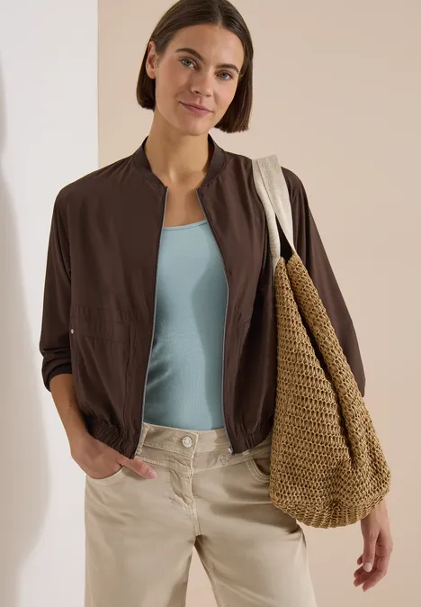 Blouson in Unifarbe mit Zipper brown cacao Frau in lässigem Outfit: hellblaues Top, braune leichte Jacke und beigefarbene Hose mit geflochtener Tasche.