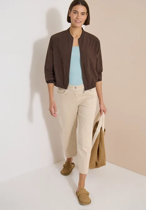 Blouson in Unifarbe mit Zipper brown cacao Frau in brauner Bomberjacke, hellblauem Top, hellen Hosen, grünen Loafers und Korbtasche vor hellem Hintergrund.