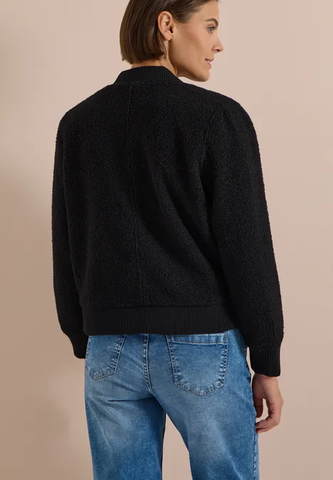 Eine Frau steht in einer schwarzen Fleecejacke und blauen Jeans, Rückenansicht vor beigem Hintergrund.