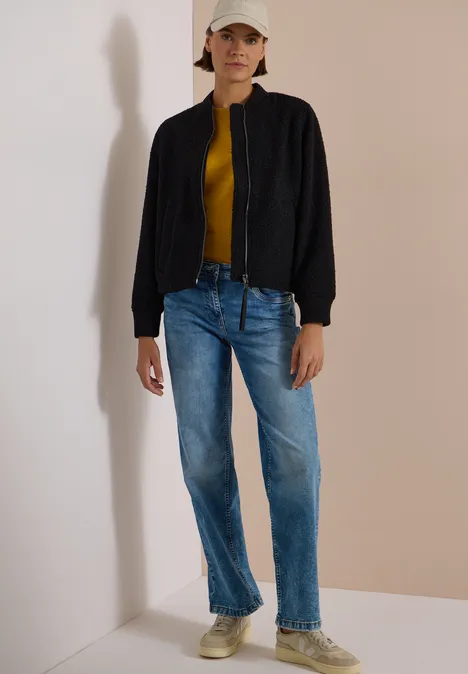 Person steht in Zimmerecke, trägt Jeans, gelbes Shirt, schwarze Jacke, Sneaker und Baseballcap.