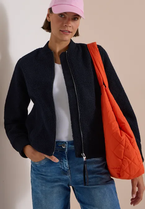 Frau trägt schwarze Bomberjacke, weißes Top, Jeans, rosa Cap und orange Stepp-Umhängetasche.