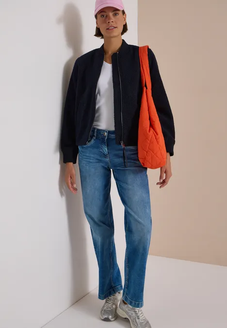 Person in lässigem Outfit: Jeans, weißes Shirt, schwarze Jacke, rosa Cap, graue Sneaker und orange Tasche vor hellem Hintergrund.