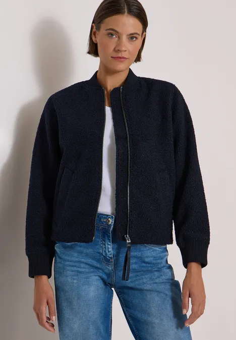 Frau in dunkelblauer Fleece-Bomberjacke, weißem Shirt und blauen Jeans vor schlichtem Hintergrund.